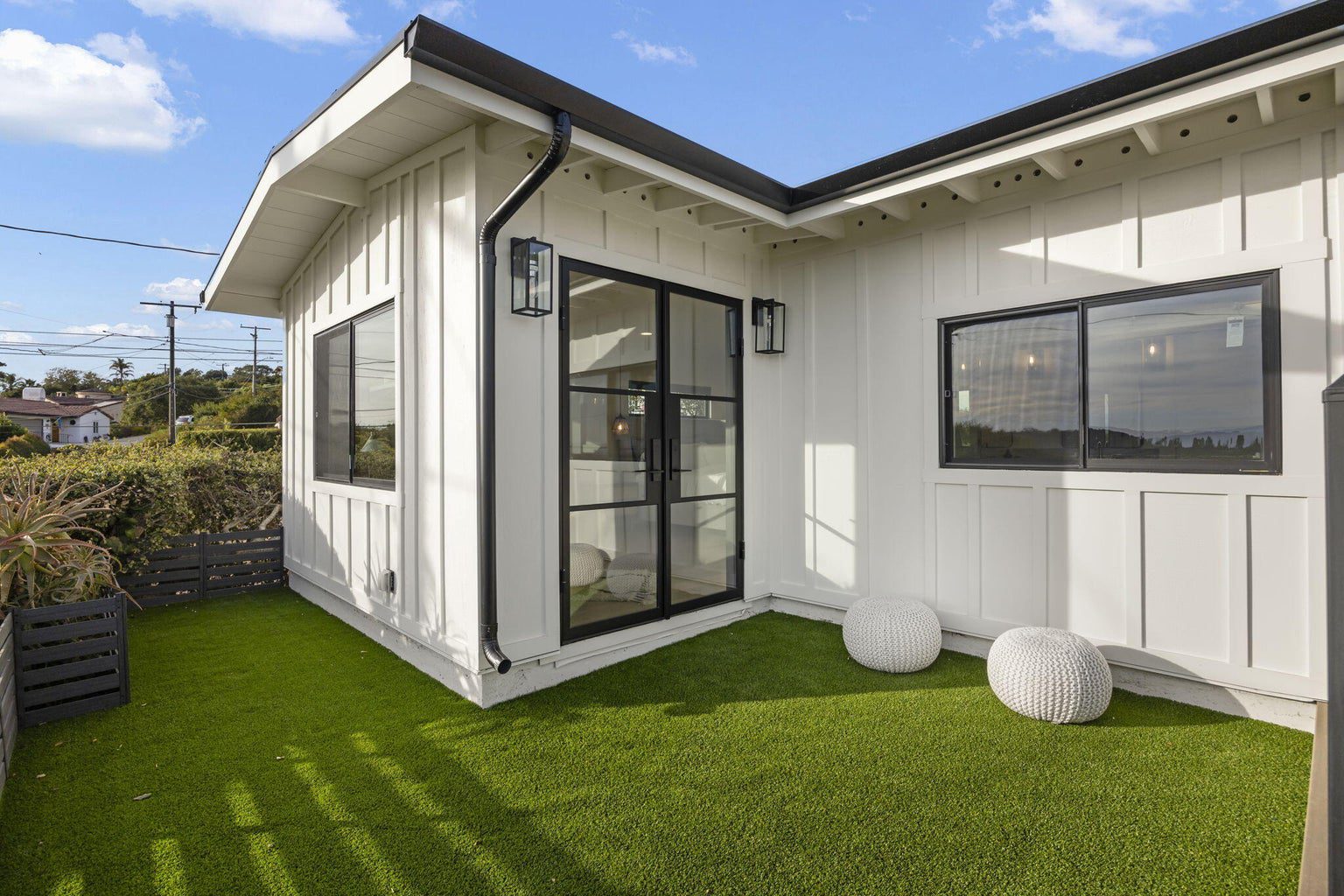 A modern white exterior house with black trim, featuring a glass door that opens to a small yard covered with artificial grass and two round white outdoor stools.